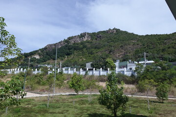 Scenic view of a green mountain with rocky outcrops behind residential houses and white fences under a bright sky in Cam Ranh, Vietnam. The foreground shows a quiet garden with young trees and trimmed