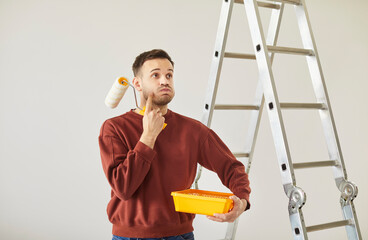 Portrait of young handsome painter man wearing casual clothes holding paint roller and tools for renovation with confused thinking face expression. Repair home renovating house concept.