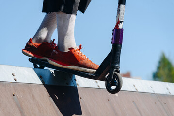 modern skate park, young boy with scooter stands on edge of skateboard ramp outdoors in city park in summer for skateboarding, close-up view of legs, roller skating and bicycle riding, extreme sport © Anton Pentegov