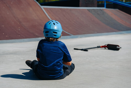 modern skate park, young boy with scooter sits near skateboard ramp outdoors in city park in summer, for skateboarding, fall or injury, roller skating and bicycle riding, extreme sport - Powered by Adobe