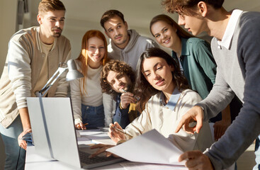 Group of young people gathered around desk in classroom, discussing project and analyzing documents. Happy university students collaborating, using laptop and sharing ideas during educational teamwork