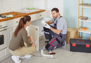 Professional male plumber trying to fix siphone in kitchen sink, explaining woman client what is wrong, trying to solve problem with pipe, cleaning out the clogged drain. Plumbing concept