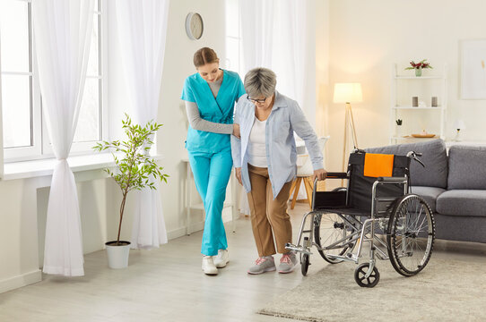 Young nurse helping senior elderly patient with disability to walk in retirement home. Physiotherapist or health care worker helping old disabled woman in wheelchair in rehab. Nursing concept.