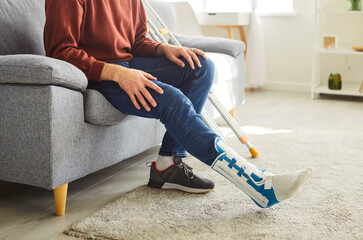 Unrecognizable faceless man with broken leg in plaster cast, posing on comfortable couch in living...