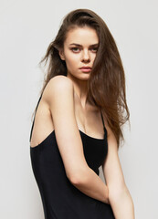 Confident young woman with long hair posing in minimalistic black outfit against a soft gray background, expressing elegance and natural beauty.