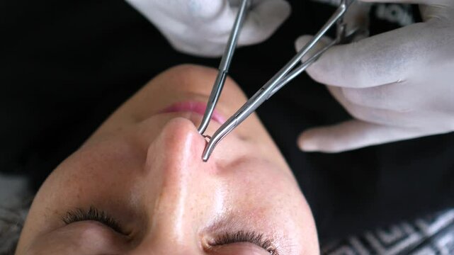 Professional piercer carefully inserting septum ring using receiving tube and forceps while client lies on treatment table, demonstrating precise body modification technique