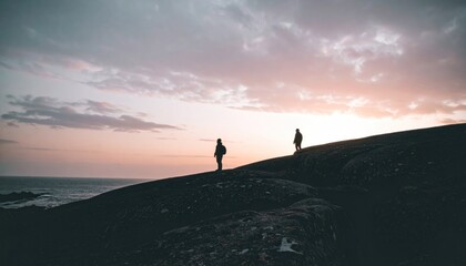 Fototapeta premium Silhouetted figures stand atop rocks at twilight; ocean, clouds, soft light