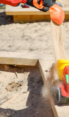 Child’s gloved hand pouring sand from a plastic shovel into a colorful toy truck in a sandbox.  Concept of childhood play, outdoor activity, motor skills, spring fun.
