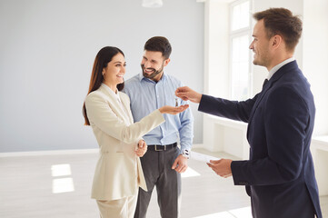 Professional realtor handing keys to smiling young woman in bright empty room while her partner...