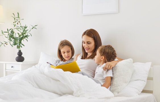 Mother lies in bed with her two children in morning and reads book looking at illustrations. Woman is reading color book aloud, while interested kids listen attentively while lying under white blanket