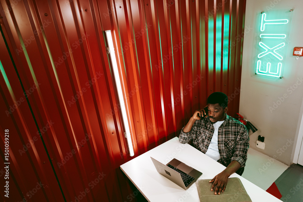 Poster working professional engaged in a phone call while using a laptop in a modern workspace with distinc