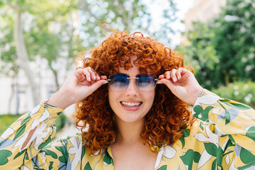 Red-haired woman wearing sunglasses standing amid verdant park greenery, her colorful floral dress...