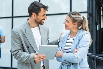 Diverse team of corporate professionals in formal attire collaborating outdoors, engaged in strategic discussion while analyzing data on laptop in front of modern glass office building.