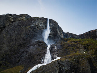 Aerial shot: Powerful waterfall with crystal clear water from a melting glacier, flowing down rocky mountain ledges.