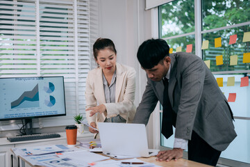 Cheerful Asian businessman and woman looking at laptop screen, analyzing and celebrating good news or project success in office.