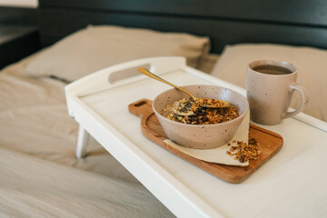 Flatlay of cozy breakfast tray with granola, milk, amber candle, and napkin on a soft beige bed....