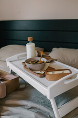 Stylish breakfast tray with a bowl of granola, milk bottle, and candle on a cozy bed, next to a beautifully wrapped gift. A calm and warm morning celebration scene