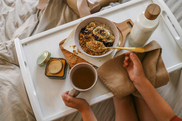 Child’s hands holding spoon and mug while enjoying breakfast in bed with granola, milk, candle, and a wrapped gift in the background. Cozy and calm morning scene