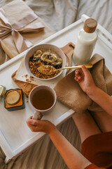 Child’s hands holding spoon and mug while enjoying breakfast in bed with granola, milk, candle, and a wrapped gift in the background. Cozy and calm morning scene