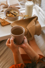Child’s hands holding spoon and mug while enjoying breakfast in bed with granola, milk, candle, and a wrapped gift in the background. Cozy and calm morning scene