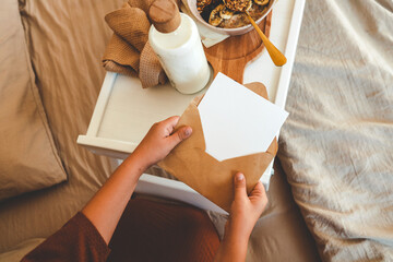 Child opening kraft envelope with card on a cozy bed, next to breakfast tray with granola and milk,...