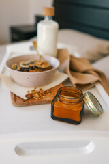 Close-up of a small amber glass jar candle with metal lid on a white tray, next to a bowl of granola with banana, dried fruits and milk in soft morning light
