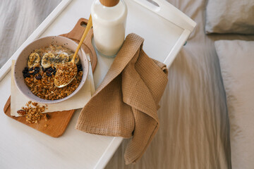 Flatlay of cozy breakfast tray with granola, milk, amber candle, and napkin on a soft beige bed....