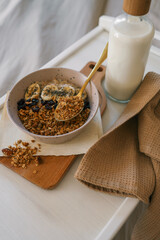 Flatlay of cozy breakfast tray with granola, milk, amber candle, and napkin on a soft beige bed....