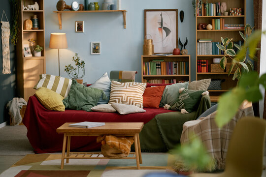 Interior of cozy living room with mix of colorful cushions, wooden bookshelves, indoor plants, cozy lighting, and eclectic decor