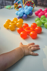 Colorful plastic sensory toys in red, yellow, green, blue, and purple lie on a white table in sunlight, with a toddler’s hand reaching up to touch them. Fun educational toys for babies or toddlers