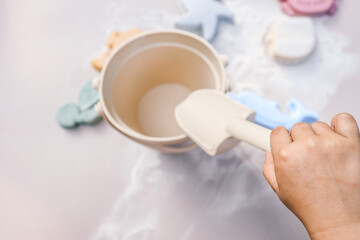 Toddler playing with pastel-colored silicone beach toys in the sand, including a bucket, scoop, starfish, dolphin, and whale shapes. Warm sunlight creates soft shadows and a peaceful summer mood