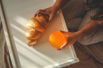 Child's hands reaching for a croissant and orange juice on a breakfast tray in bed, soft sunlight...
