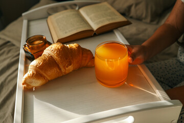 Child's hands reaching for a croissant and orange juice on a breakfast tray in bed, soft sunlight...