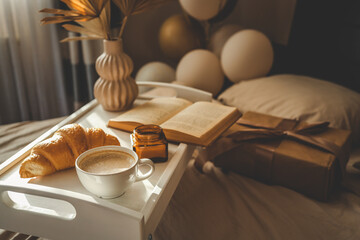 Elegant morning scene with coffee, croissant, book, gift box, dried palm leaves, and balloons on a...