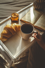 Breakfast in bed with croissant, coffee, candle, and open book on a white tray, lit by soft morning light. A child’s hand holds the cup, adding warmth and intimacy to the scene