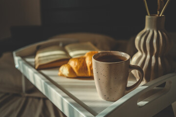 Beautiful morning scene with a croissant, candle, coffee, open book, and decorative vase on a white tray in bed, softly lit by natural sunlight through curtains