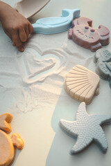 Child’s hand drawing a heart in fine white sand, surrounded by pastel sea-themed toys including a...