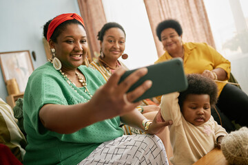 Group of smiling Black family with baby and senior woman taking selfie using smartphone gathering in cozy living room