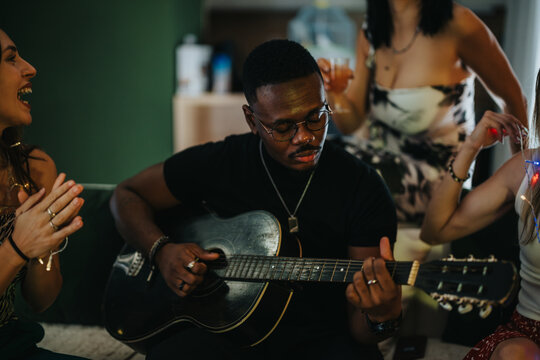A man playing guitar while friends clap and enjoy a lively atmosphere at a cheerful gathering, suggesting fun and community around music and happiness.