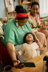 Vertical shot of happy Black woman snapping selfie with cheerful little daughter while spending family time at in living room