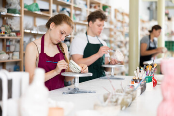 Girl teenager student in apron trained to paint ceramic cup in workshop