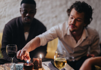 Two friends sit in a cozy setting sharing drinks while one lights a candle, creating a warm and tranquil atmosphere. Glasses of wine add a touch of sophistication to the scene.