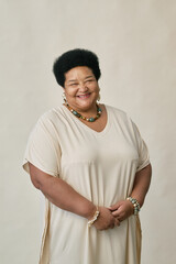 Vertical portrait of Black woman standing against neutral background with relaxed smile and hands clasped while wearing beaded necklace and bracelet posing in studio