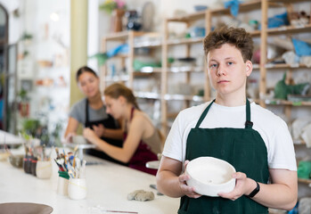 Guy teenager student in apron posing with ceramic bowl in his hands in ceramic workshop