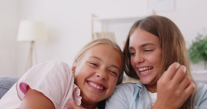 Close up of two happy funny young girls sitting on sofa, having fun and fooling around. Cheerful playful sisters or friends laughing, tickling each other and playing carefree in living room at home.