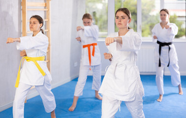 Motivated sporty teenage girl with group of young female karate practitioners wearing white kimonos diligently performing kata routines to hone martial arts skills in training room..