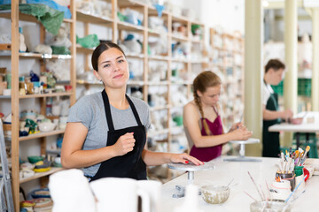 Young woman teacher in apron posing against backdrop of ceramic workshop