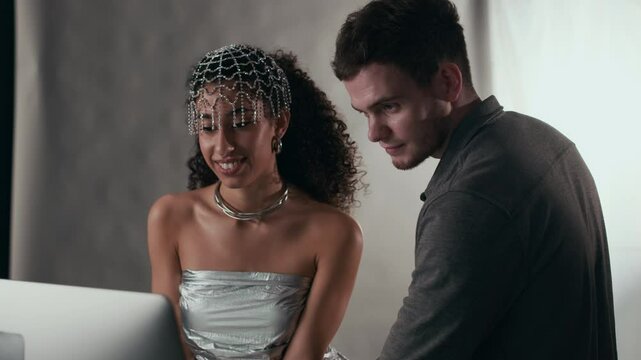 Medium closeup of young beautiful biracial model in silver designer dress looking at PC screen with Caucasian male photographer and laughing while discussing pictures made in professional photo studio
