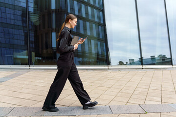 Professional woman walking confidently outside a modern building while using a tablet in an urban...