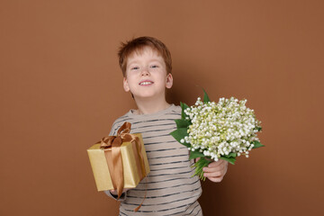 Boy with bouquet of beautiful lily-of-the-valley flowers and gift on brown background